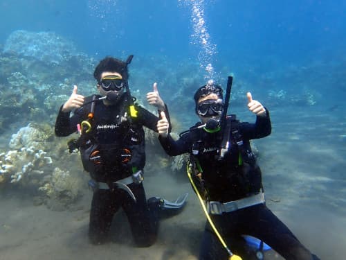 水中で砂地に座ってサンゴ礁をバックに記念撮影する2名のダイバー