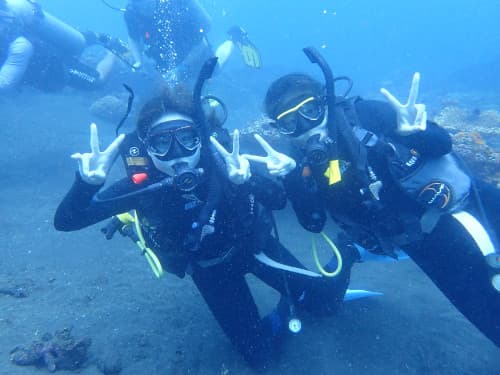 砂の水底に座ってピースサインをする２人の女性ダイバー