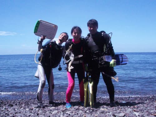 海をバックにビーチに立って記念撮影するダイビング器材を背負ったウェットスーツ姿の3名のダイバー