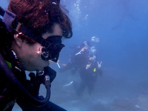バリ島のキレイな海を新鮮に感じている体験ダイビングのお客様の横顔とその向こうに見えるピースしてる体験ダイバー