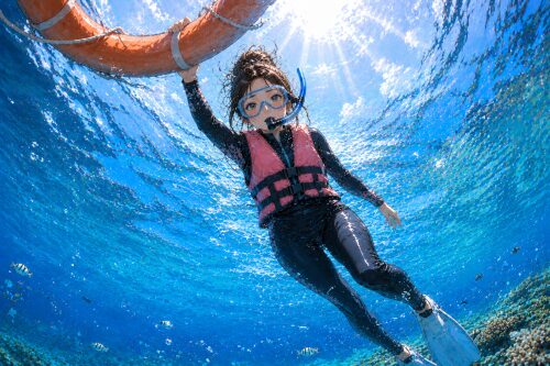 水中から見上げた構図で、フロートに片手で掴まりライフジャケットを着て浮かぶ女性シュノーケラー